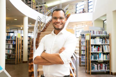 Photo of a man in a library who is smiling with his arms crossed